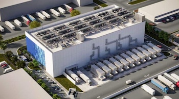 Vista aérea de un gran centro de datos blanco con una fachada azul, unidades de refrigeración en el techo, bahías de carga y camiones, rodeado de carreteras y vegetación