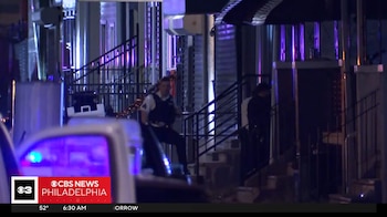 Un oficial de policía con chaleco antibalas permanece junto a las escaleras de un inmueble iluminado por luces de patrulla en la noche