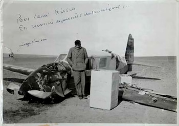 Hombre con abrigo y gorra de pie junto a avión con la parte delantera destrozada en un desierto. Foto en blanco y negro del accidente