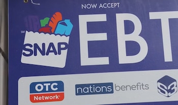 Primer plano de un cartel azul que dice "NOW ACCEPT EBT SNAP" con el logo de una bolsa de compras. También se ven los logos de OTC Network, Nations Benefits y S3