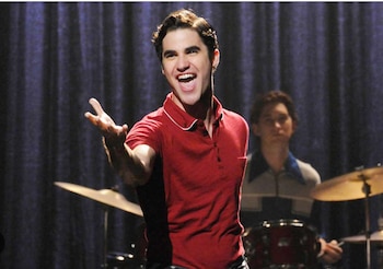 Blaine Anderson, un hombre con camisa polo roja y el pelo peinado, canta con la boca abierta y el brazo extendido, con un baterista detrás