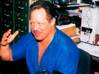 Hombre de mediana edad con cabello oscuro y bigote, camisa azul abierta, gesticula con la mano izquierda, sentado frente a un ordenador y un teclado en un interior