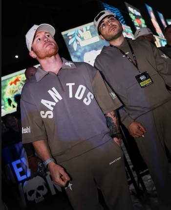 Saúl Canelo Álvarez y otro hombre con conjuntos deportivos grises y gorras blancas miran hacia arriba en un espacio interior