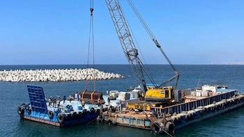 Una grúa amarilla en una barcaza oscura levanta un gran bloque de una barcaza azul con personas. Al fondo, un rompeolas de tetrapodos y el mar