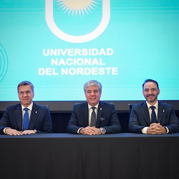 Tres hombres con traje y corbata sentados frente a una mesa negra con el logo y el nombre de la Universidad Nacional del Nordeste en el fondo