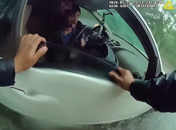 Primer plano de dos manos intentando abrir la puerta de un coche blanco sumergido en agua. Dentro del coche, una persona se ve sentada al volante, con el rostro pixelado