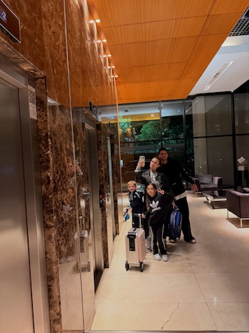 Una mujer, un hombre y dos niños posan con tres maletas frente a puertas de ascensor y un espejo en el lobby de un edificio
