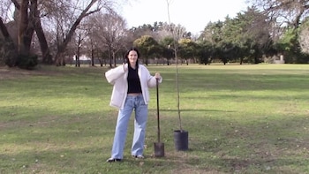 Mujer de cabello oscuro con chaqueta blanca y jeans azules sostiene una pala, parada junto a un árbol joven en maceta negra en un parque verde