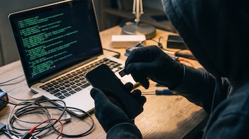 Una persona encapuchada con guantes negros inserta una tarjeta SIM en un teléfono móvil, junto a una laptop con código de programación y cables en una mesa.