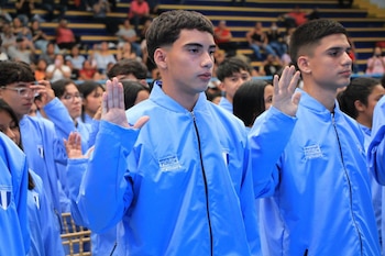 Jóvenes con chaquetas deportivas azules levantando la mano derecha en un recinto con graderías al fondo