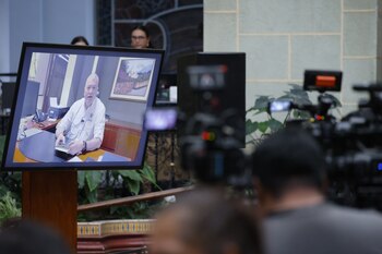 Un monitor de pantalla plana muestra a un hombre sentado tras un escritorio, rodeado por personas y cámaras de televisión