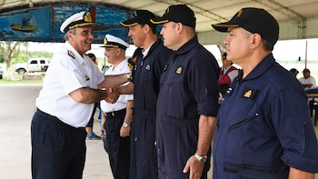 Varios hombres en uniformes navales se encuentran al aire libre; uno de camisa blanca y gorra saluda con la mano a otro de mono azul oscuro y gorra