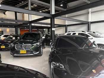 Interior de un concesionario. Varios automóviles estacionados, incluyendo un Porsche oscuro en primer plano, un BMW verde y un Mazda oscuro. Techo con vigas metálicas