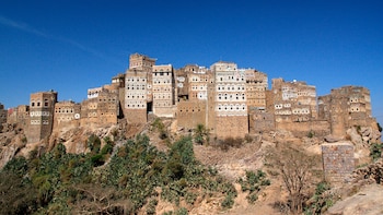 Edificios altos de piedra con fachadas decoradas construidos sobre un acantilado rocoso bajo un cielo azul despejado