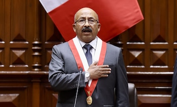 Hombre calvo con gafas, bigote, terno gris, corbata azul, banda presidencial bicolor y medalla, con la mano en el pecho frente a un micrófono y bandera