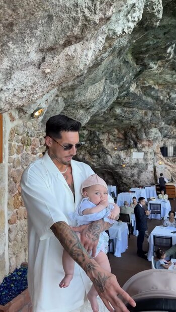 José Sosa, con camisa blanca y tatuajes en los brazos, sostiene a un bebé con un gorro rosa. Al fondo, un restaurante con paredes rocosas y mesas
