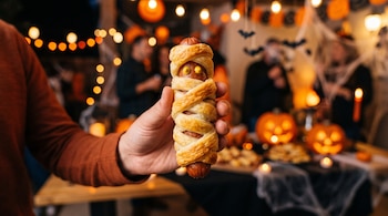 Una mano sostiene una salchicha envuelta en tiras de masa con dos puntos de mostaza, frente a calabazas talladas y luces.