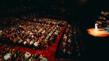 Vista general de la sala de un teatro llena de espectadores y un artista sobre el escenario bajo un foco de luz