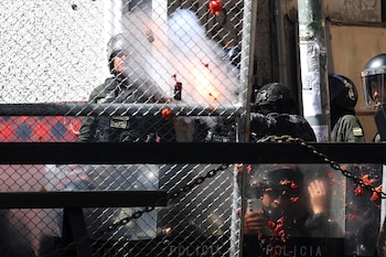 Agentes de policía con equipo antidisturbios se ven parcialmente detrás de una valla metálica. Humo y escombros rojos brillantes flotan en el aire, con un escudo cubierto de manchas rojas