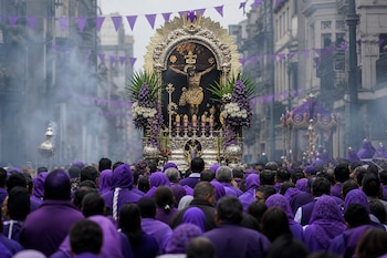 Multitud de espaldas con vestimenta morada frente al anda procesional de Cristo crucificado, rodeada de flores, velas e incienso en una calle.