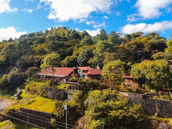 Vista aérea de casas de techo rojo en una ladera boscosa con vegetación exuberante, cielo azul y nubes. Una tabla de surf azul y blanca y motos se ven abajo