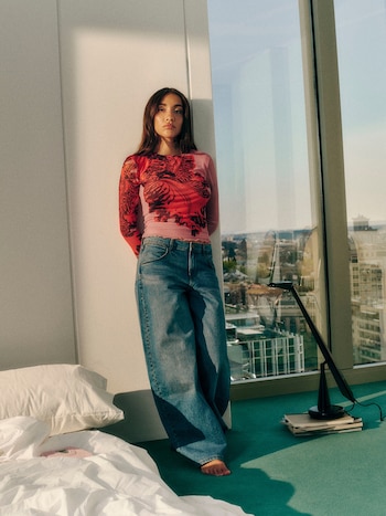Maria Becerra, descalza, con top rosa y rojo estampado y jeans anchos. De pie junto a una pared blanca y un ventanal con vista a la ciudad. Cama y lámpara de mesa
