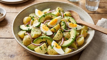 Cuenco con ensalada de papas, rodajas de palta, mitades de huevo cocido, aros de cebolla morada, perejil picado, pimienta y cuchara de madera.