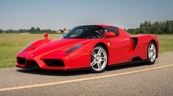 Un Ferrari Enzo rojo brillante estacionado en una carretera asfaltada con una línea amarilla, con campos verdes y árboles en el fondo bajo un cielo azul.