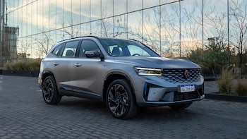 Un automóvil SUV Renault Koleos de color gris plateado está estacionado sobre adoquines frente a un edificio con fachada de vidrio