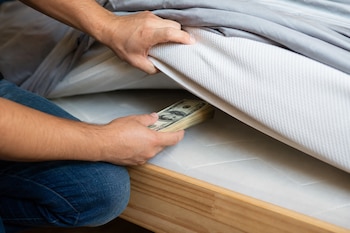 Unas manos levantan la esquina de un colchón en una cama de madera y colocan debajo un fajo de billetes de dólar.
