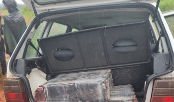 Baúl de un coche blanco abierto mostrando grandes paquetes rectangulares envueltos en plástico transparente, presumiblemente droga, bajo la lluvia
