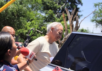 Hombre de cabello blanco junto a un vehículo, rodeado por personas con micrófonos. Un periodista sostiene un micrófono rojo con logo. Fondo de árboles verdes