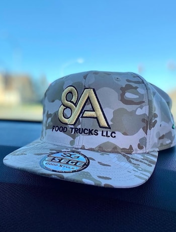 Gorra con estampado de camuflaje beige y marrón, con bordado frontal que dice 8A FOOD TRUCKS LLC y una etiqueta redonda en la visera