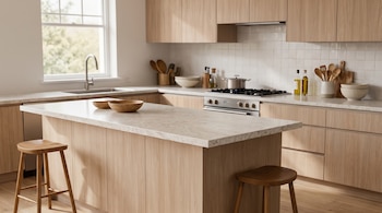 Cocina de madera clara con una isla central de encimera blanca, dos taburetes, un fregadero junto a una ventana y utensilios sobre el mostrador.