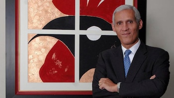 Hombre de cabello gris y traje oscuro con corbata azul, de pie con los brazos cruzados, frente a un cuadro con patrones geométricos rojos, negros, blancos y dorados