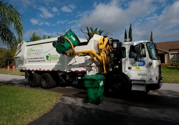 Un camión de basura blanco de Miami-Dade con un brazo amarillo levanta un contenedor verde. Otro contenedor verde está en la acera; se ven palmeras y una casa