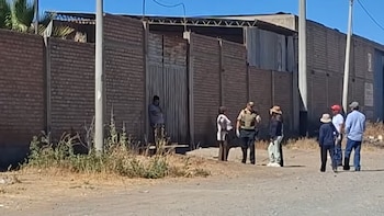 Un agente policial y siete personas en una calle de tierra frente a un muro de ladrillos, una puerta de metal y un edificio industrial bajo un cielo azul claro
