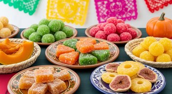 Mesa con surtido de dulces cristalizados mexicanos en platos y cestas. Bolas, cubos, anillos de piña, higos, calabaza cocida y papel picado al fondo.