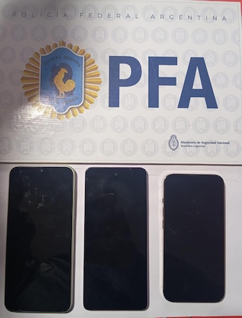 Una vista aérea de tres smartphones con pantallas oscuras sobre un documento blanco con el logo de la Policía Federal Argentina (PFA) y el escudo nacional