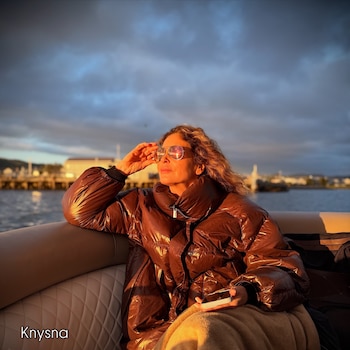 Una mujer con chaqueta acolchada marrón y gafas de sol sentada en un barco, sosteniendo un teléfono, con el sol bajo reflejado y un cielo nublado