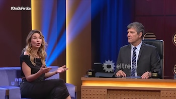 Marcela Kloosterboer, de cabello rubio ondulado y vestido negro, sentada en un sofá azul, gesticula mientras habla con un hombre en un set de televisión iluminado