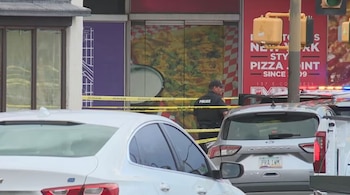 Un oficial de policía se encuentra detrás de una cinta amarilla de seguridad en la calle. Varios vehículos y una tienda de pizza son visibles