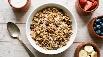 Fotografía cenital de un bowl blanco con granola, nueces, semillas, pasas y coco, junto a una cuchara, yogur, frutillas, arándanos y banana en una mesa de madera.