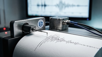Aguja metálica de sismógrafo que traza líneas sobre papel blanco junto a un sensor de metal y una pantalla con ondas al fondo.
