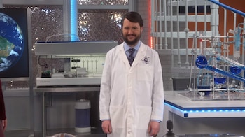 Un hombre con barba y bata blanca sonríe en un laboratorio. Se ven matraces con líquidos azules, un sistema de destilación y una pantalla con la Tierra
