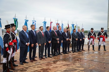 Once hombres de traje y corbata se alinean junto a dos soldados con uniforme de gala frente a una fila de banderas y astas en un patio mojado