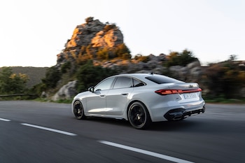Automóvil Audi A5 blanco en movimiento con matrícula alemana, visto desde la parte trasera izquierda. Carretera con línea discontinua blanca. Montañas y árboles de fondo borroso