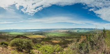 Vegetación verde, arbustos secos, campos, colinas, un cuerpo de agua, montañas distantes y cielo azul con nubes blancas en una vista panorámica