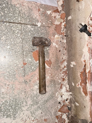 Un mazo de hierro y mango de madera yace sobre un suelo gris salpicado con escombros de ladrillo rojo y yeso blanco, junto a un enchufe negro