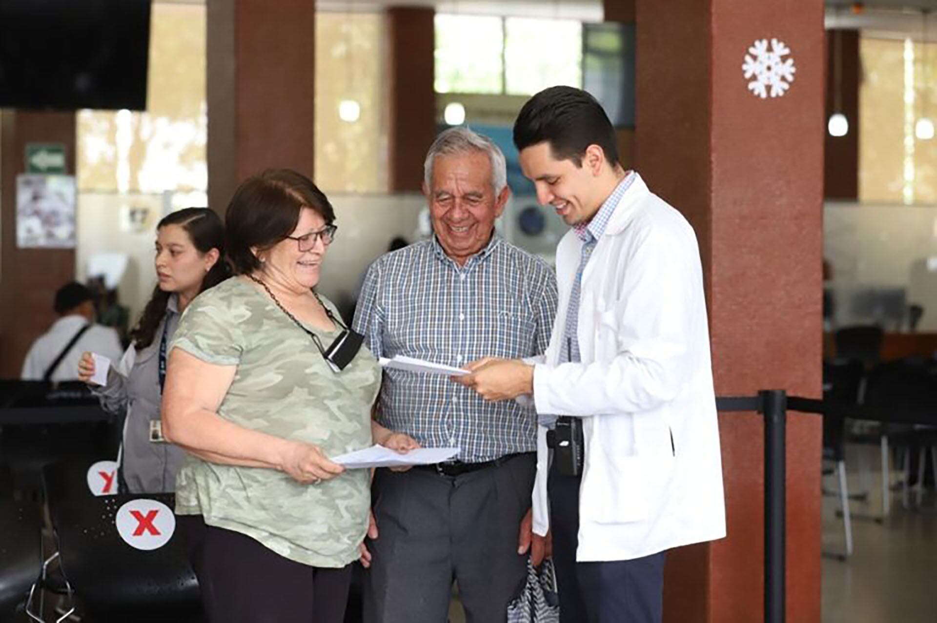 Un joven profesional brinda asistencia y orientación a una pareja de adultos mayores en lo que parece ser un vestíbulo o área de atención en un centro de eventos. (redes IGSS)